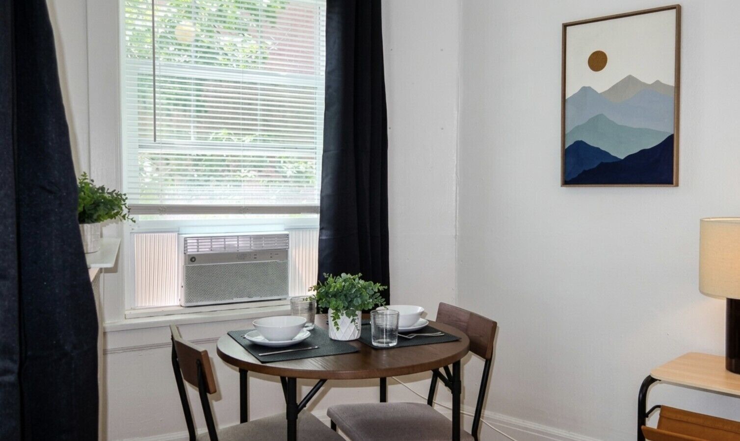 A dining area with a table set for two, a potted plant, and a wall-mounted painting.