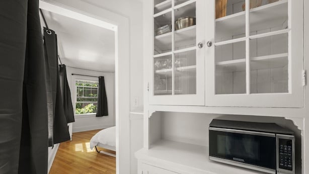 Une pièce comprenant un lit, une fenêtre avec des rideaux, une armoire blanche avec un four à micro-ondes, et une porte donnant sur une autre pièce.