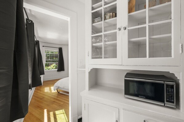 A room with a bed, a window with curtains, a white cabinet with a microwave, and a door leading to another room.