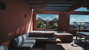 A sunlit outdoor seating area with a sofa, a table, and a view of palm trees and mountains.