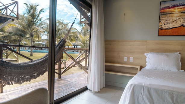 A bedroom with a large window, a hammock, a bed, and a view of a pool and palm trees.