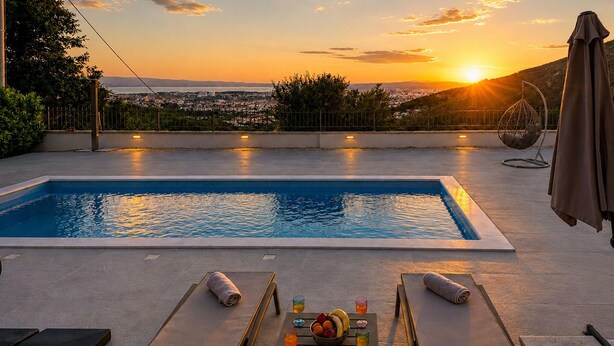 Piscina rectangular con vistas al atardecer, sillas de descanso, una mesa con bebidas y fruta, y una zona cubierta con una silla.