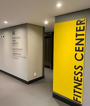 Corredor com parede onde se destaca um grande letreiro amarelo com a inscrição 'FITNESS CENTER' e parede branca com borda preta e a letra 'M' no topo.