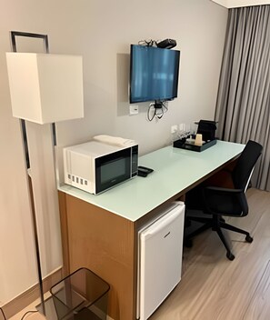 Quarto de hotel com escrivaninha, micro-ondas, geladeira, cadeira de escritório preta, TV fixada na parede e luminária de chão.