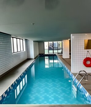 Una piscina interior con agua cristalina, paredes de azulejos y ventanas que dejan entrar luz natural.