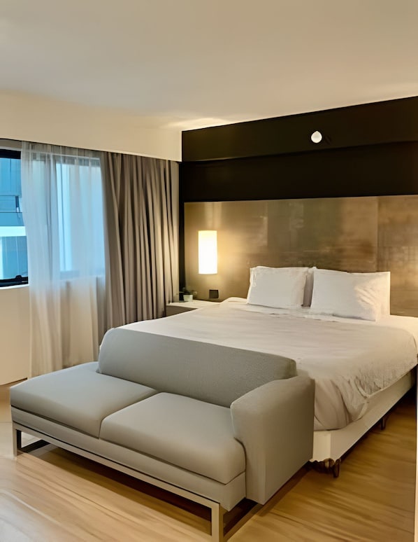 Quarto de hotel com cama, sofá, mesa de cabeceira, abajur e janela com cortinas.