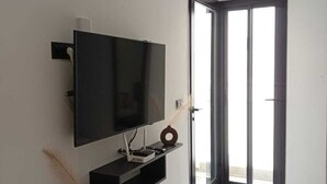 A modern living room with a flat-screen TV, a small shelf, and a sliding glass door leading to a balcony. - sandra 12 (la soukra)