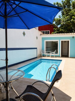 Ein Swimmingpool mit klarem, blauem Wasser, umgeben von einer gefliesten Terrasse, und ein blauer Sonnenschirm, der Schatten spendet.