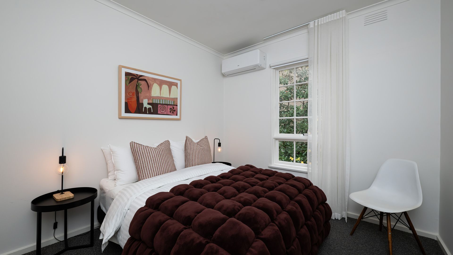 Une chambre avec un grand lit, une table de chevet avec une lampe, une chaise, une fenêtre avec des rideaux et un appareil de climatisation.