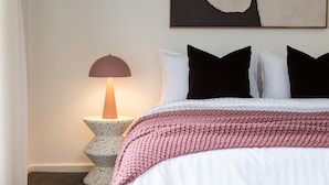 Un lit bien fait, recouvert d’une couverture tricotée rose, agrémenté d’un lampadaire moderne et d’une simple décoration murale.
