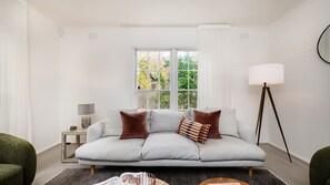 A modern living room with a grey sofa, a floor lamp, and a window with a view of trees. - Edenbrook in Prahran (Prahran)