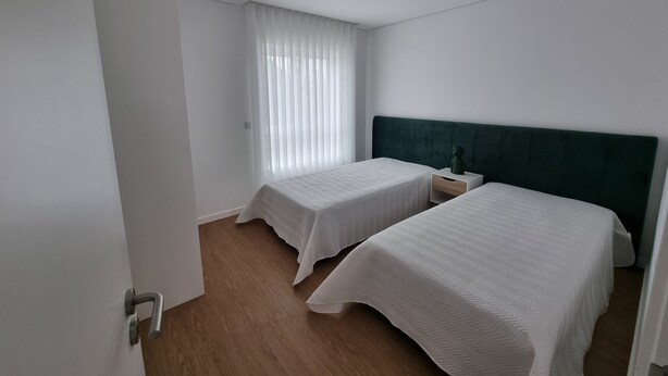 Hotellihuone, jossa on kaksi erillistä sänkyä, puulattia, vihreä pääty ja ikkuna, jossa on läpikuultavat verhot.