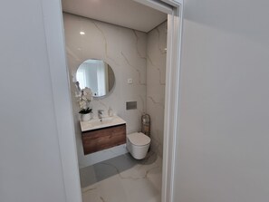 A bathroom with a marble wall, a round mirror, a white sink, a wooden vanity, and a toilet.