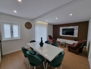Uma sala de estar moderna com uma mesa de jantar branca, cadeiras verdes, um sofá de couro, uma TV na parede e uma janela grande com cortinas.