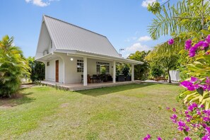 Ein Haus mit überdachter Terrasse, Rasen und tropischen Pflanzen.