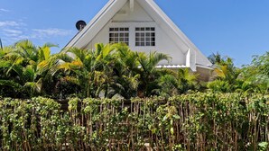 A white building with a triangular roof and multiple windows, surrounded by lush greenery.