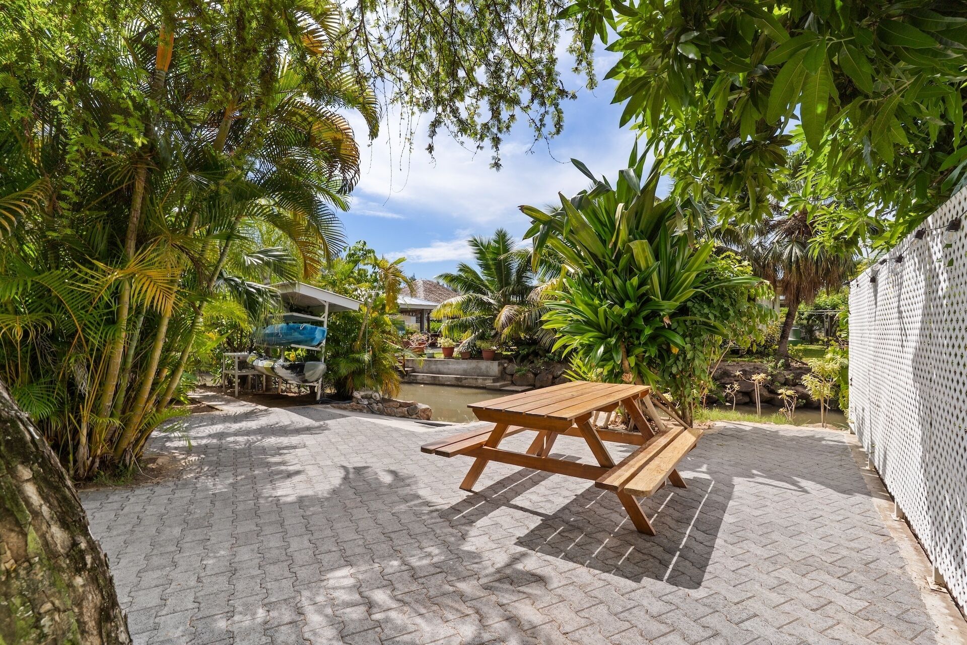 A wooden picnic table in a garden with a paved walkway, surrounded by lush greenery and a small pond.