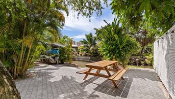 A wooden picnic table in a garden with a paved walkway, surrounded by lush greenery and a small pond.