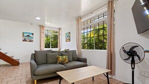 A living room with a sofa, a coffee table, a fan, and large windows with curtains.
