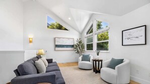 A modern living room with a grey sofa, white armchairs, a small side table, and a large window with a view of greenery. - Harbor Home - Walking Distance to Twin Lakes Beach! (Santa Cruz)