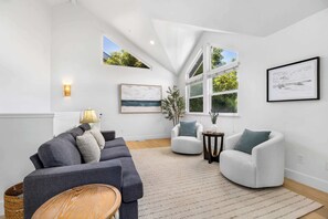 A modern living room with a grey sofa, white armchairs, a small side table, and a large window with a view of greenery. - Harbor Home - Walking Distance to Twin Lakes Beach! (Santa Cruz)