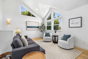 A modern living room with a grey sectional sofa, white armchairs, a small side table, and a large window with a view of greenery. - Harbor Home - Walking Distance to Twin Lakes Beach! (Santa Cruz)
