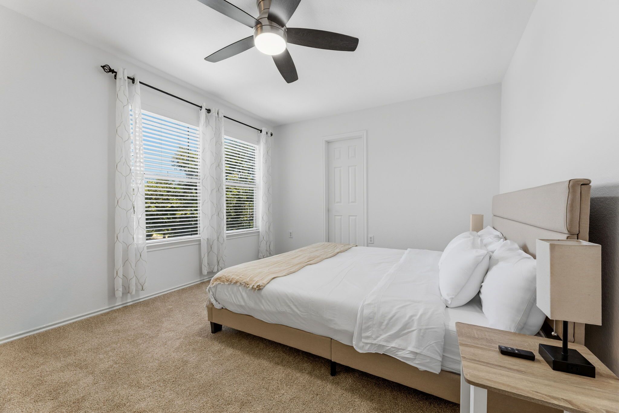 A bedroom with a bed, a ceiling fan, a nightstand with a lamp, and a window with blinds.