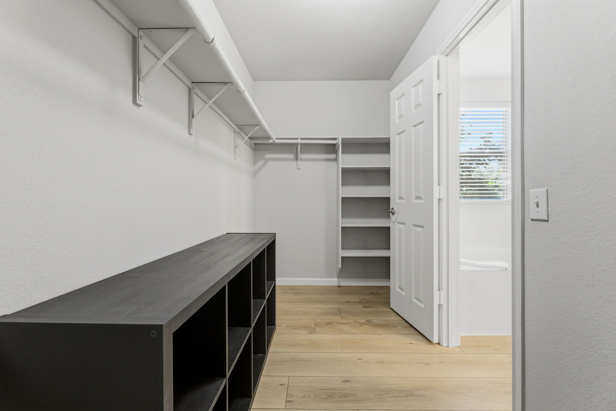 A narrow hallway with a wooden bench, white closet with shelves, and a door leading to another room.