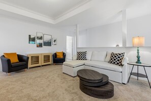 Ein Wohnzimmer mit einem Ecksofa, einem schwarzen Sessel, einem Holzschrank, einem runden Couchtisch und einer Lampe.