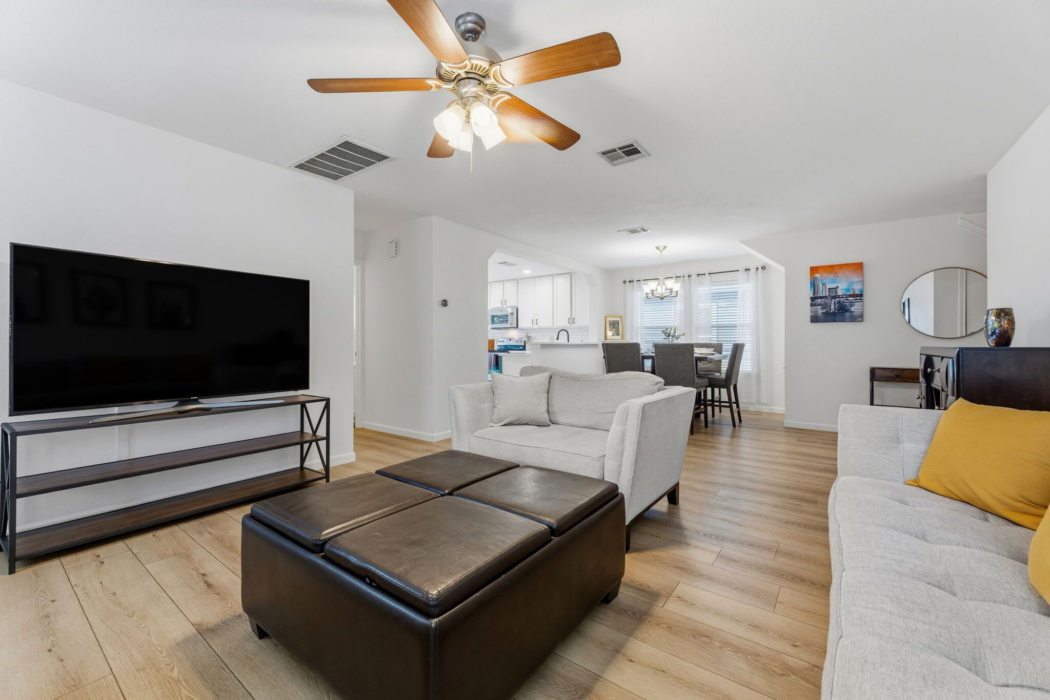 Un salon moderne doté d’un téléviseur à écran plat, d’un ventilateur de plafond, d’un canapé, d’un pouf et d’un coin repas à l’arrière-plan.