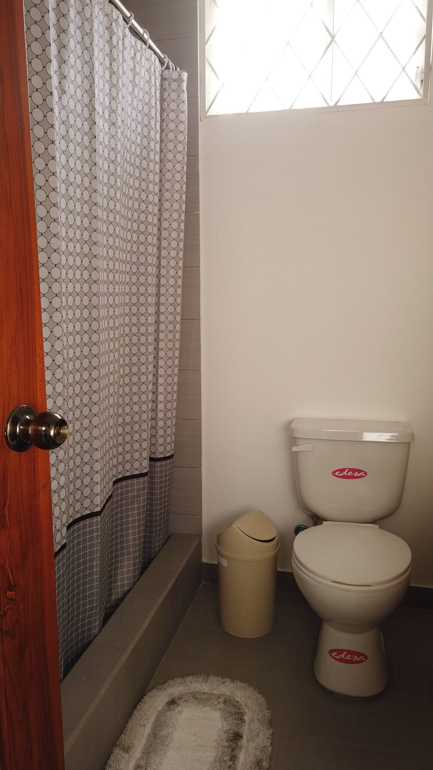 A bathroom with a toilet, a trash bin, a patterned shower curtain, and a wooden door.