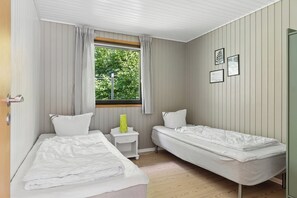 Ein Zimmer mit zwei Betten, einem Fenster mit Vorhängen, einem Nachttisch mit Lampe und gerahmten Bildern an der Wand.