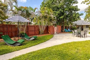 Ein Garten mit Beton-Terrasse, Rasen, zwei Liegestühlen, einem gestreiften Sonnenschirm und einem Holzzaun.