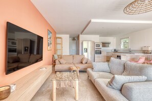 Ein modernes Wohnzimmer mit einem großen Flachbildfernseher, zwei Sofas, einem Glastisch und einer Küche im Hintergrund.