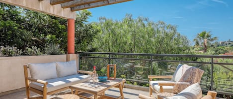 A patio with wooden furniture, a glass balustrade, and a view of greenery.
