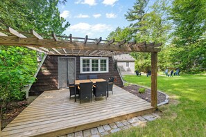 Um deck de madeira com pérgola, um pequeno edifício com janela e mobiliário de exterior.