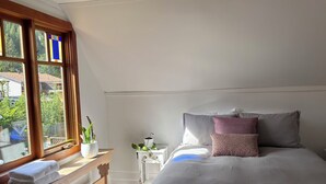 Ein Schlafzimmer mit einem Bett, Kissen, einem Nachttisch, einem Fenster mit Vorhängen und Blick ins Freie.