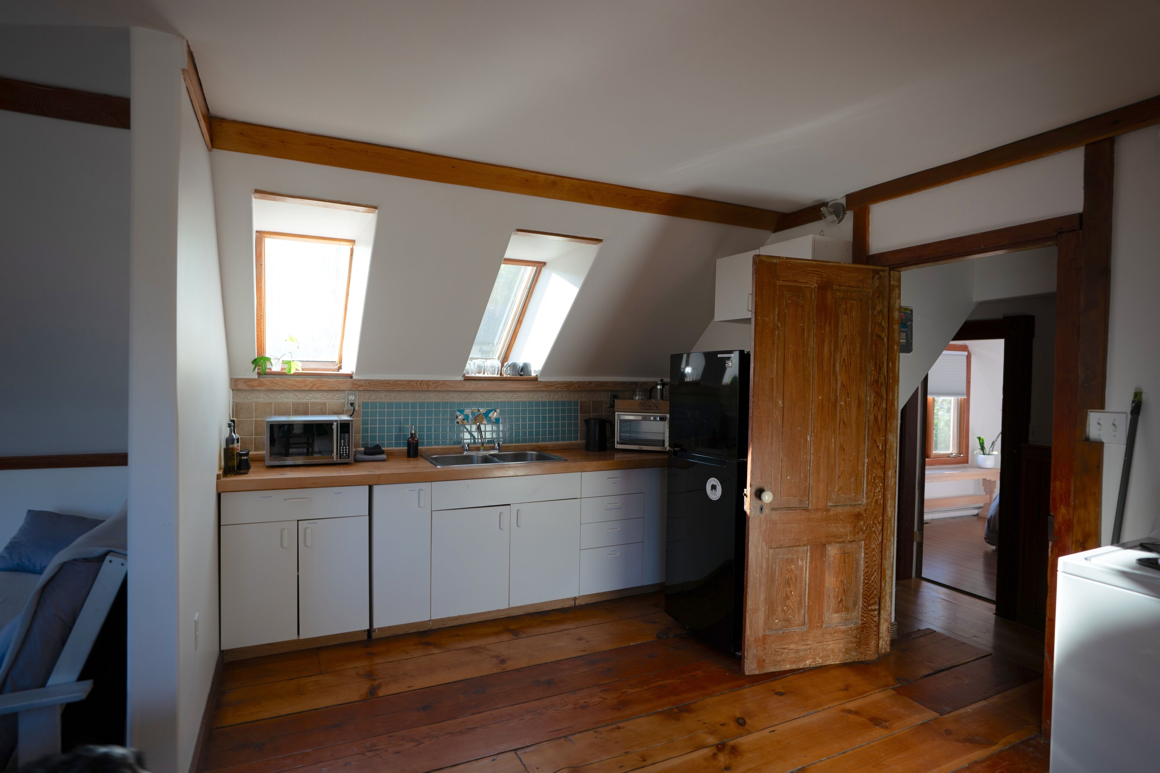 A compact kitchen with a wooden floor, white cabinets, a microwave, a toaster, and a refrigerator. There are two skylights providing natural light.