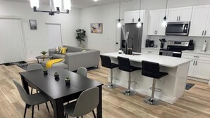 Eine moderne Küche mit weißen Schränken, einer zentralen Kochinsel inklusive Barhocker, einem Esstisch mit Stühlen und einem grauen Sofa im Wohnbereich.