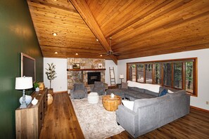 Un salon spacieux doté d’une cheminée en pierre, d’un plafond en bois et de grandes fenêtres offrant une vue sur l’extérieur.