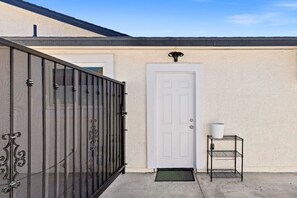 Une maison avec une porte blanche, un portail en métal noir et une petite étagère en métal sur laquelle se trouve un récipient blanc.
