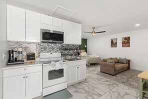 A modern kitchen with white cabinets, a microwave, and a stove. There is a dining area with a brown sofa and a coffee machine. - Brighthill Haven | Cozy Studio | 10 Min to Strip (Las Vegas)