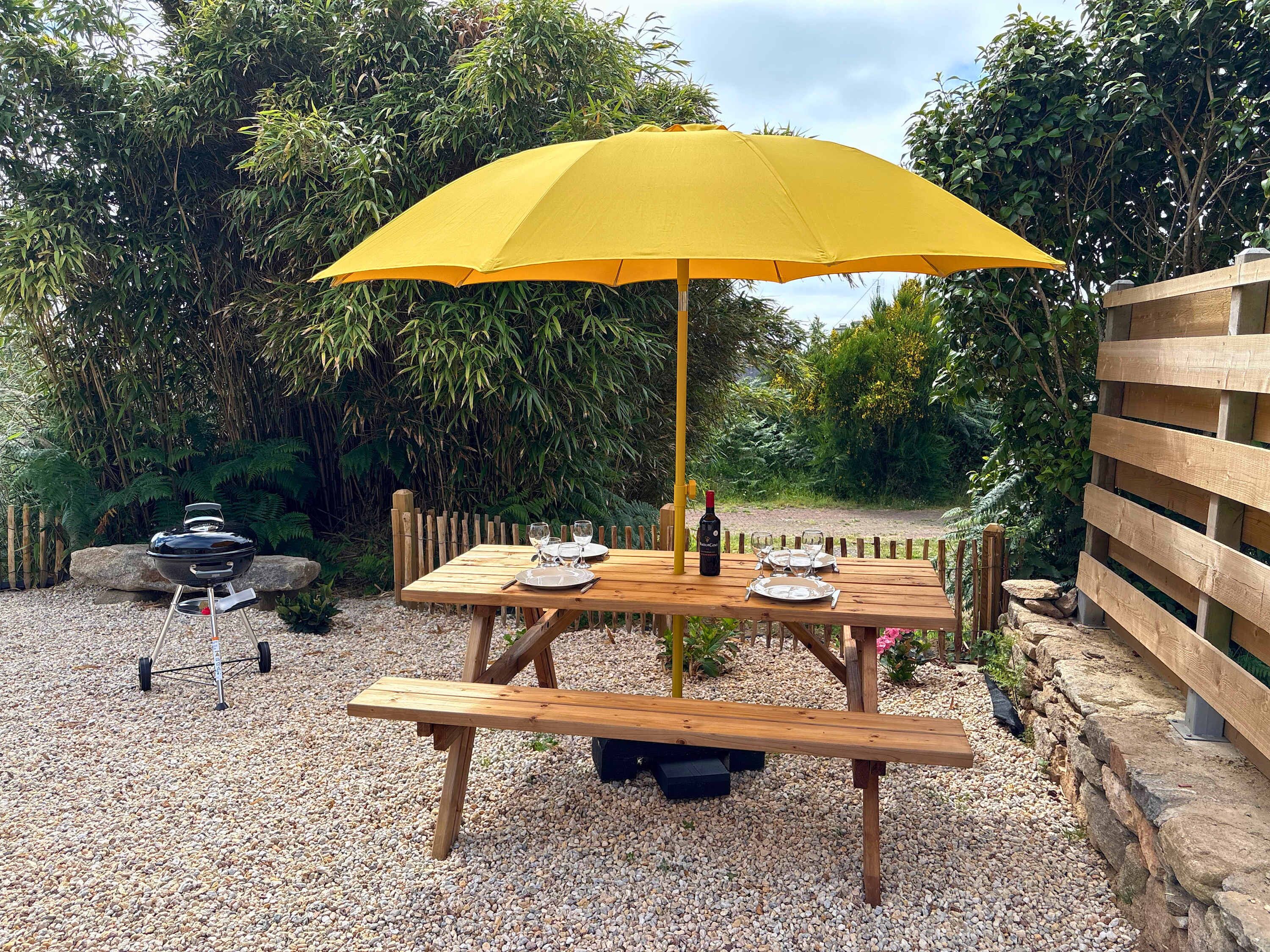 Un espace extérieur aménagé pour les repas, avec une table et un banc en bois, un parasol jaune et un barbecue.
