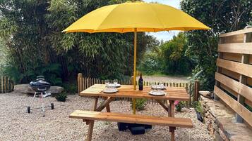 Un espace extérieur aménagé pour les repas, avec une table et un banc en bois, un parasol jaune et un barbecue.