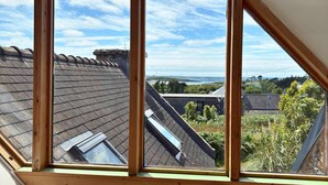 Una habitación con techo inclinado, vigas de madera y ventana con vistas a tejados y vegetación.