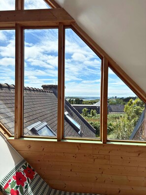 Ein Zimmer mit schrägen Wänden, Holzbalken und einem Fenster mit Blick auf Dächer und Grünflächen.