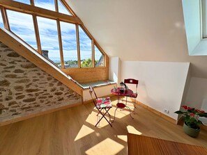 Ein Schlafzimmer im Dachgeschoss mit schrägem Dach, einer Holztreppe, einem kleinen Tisch mit Stühlen und Blick auf Himmel und Landschaft.