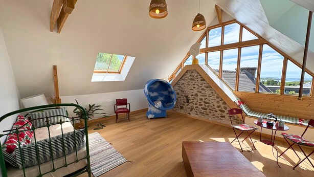 Una habitación estilo loft con techo inclinado, vigas de madera, un ventanal grande, una pared de piedra, una cama de metal verde, una mesa de madera y una silla azul esférica.