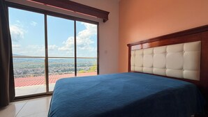 A bedroom with a large window, a bed, and a view of a cityscape.