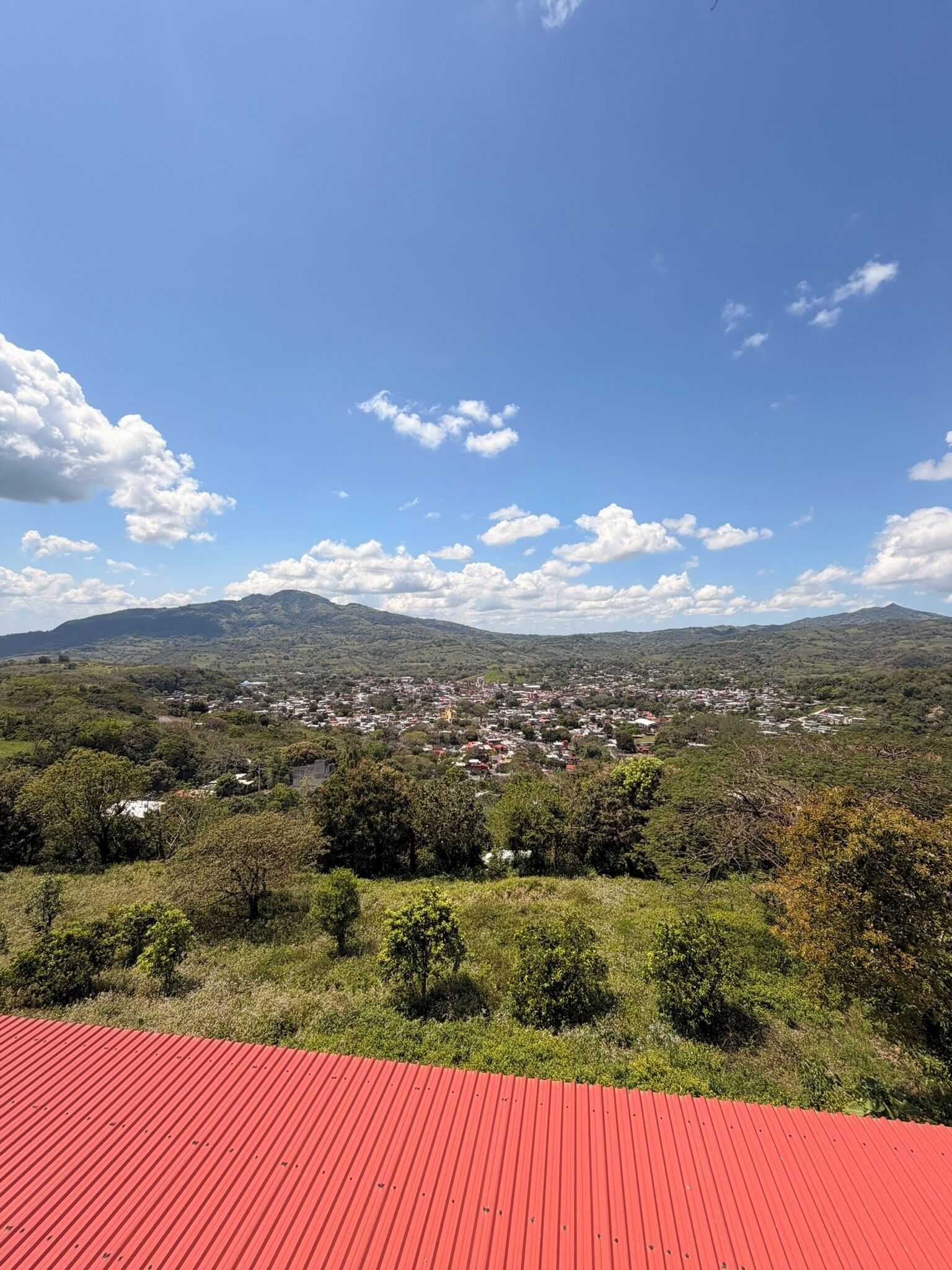 A panoramic view of a town with a red-roofed building in the foreground, surrounded by green hills and a clear blue sky.